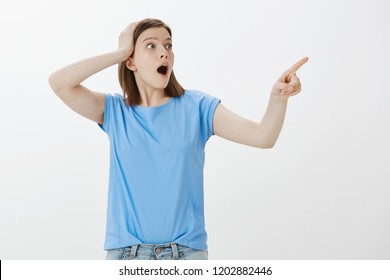 No Way, Look At This Large Wave. Impressed Shocked Attractive European Woman In Blue T-shirt, Holding Hand On Head And Pointing Right, Dropping Jaw From Surprised And Amazement Over Grey Wall