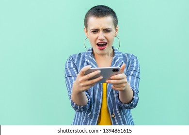 No way! Portrait of beautiful unbelievable with short hair young woman in striped suit standing, playing game on her phone with shocked face. Indoor, isolated, studio shot, green background