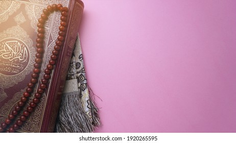 The Noble Quran with tasbih or rosary beads and prayer mat over pink background. Arabic word translation "The Noble Al Quran (Holy book of Muslims)". Ramadan Conceptual photo