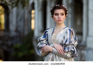 Noble Young Woman With Elegant Hairstyle In Medieval Dress In The Garden Of Old Castle