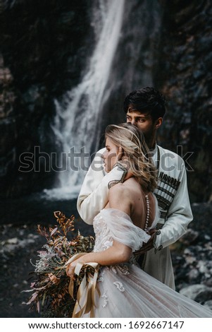noise effect, selective focus: incredibly enamored brides hugging, kissing and posing for a photo on the incredible rocky mountains background with a big waterfall. Wedding in Georgia.