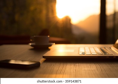 Nomad Work Concept Image Computer Coffee Mug and Telephone on black wood Table and Evening Sunlight shining throw large windows focus on Edge of Laptop
