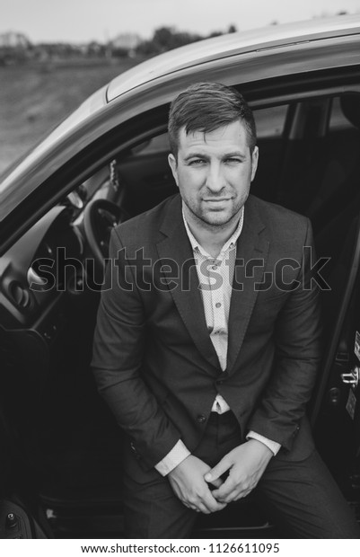Non beard.\
Man in a business suit is sitting in the car with open door.\
Businessman posing on camera outdoors. Luxury Life. Lifestyle\
photo. Ideal for commercial and phone\
photo.