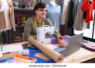 Non binary man tailor using sewing machine and laptop at atelier