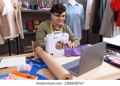 Non binary man tailor using sewing machine and laptop at atelier
