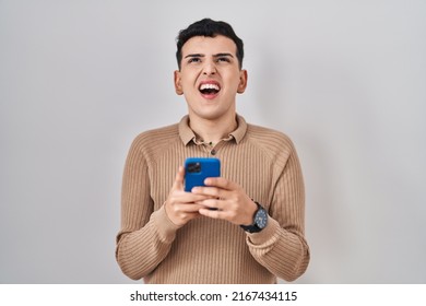 Non Binary Person Using Smartphone Typing Message Angry And Mad Screaming Frustrated And Furious, Shouting With Anger Looking Up. 