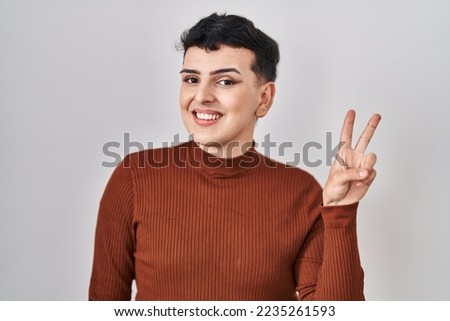 Non binary person wearing make up standing over isolated background smiling with happy face winking at the camera doing victory sign. number two. 