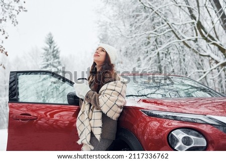 Non urban scene. Beautiful young woman is outdoors near her red automobile at winter time.