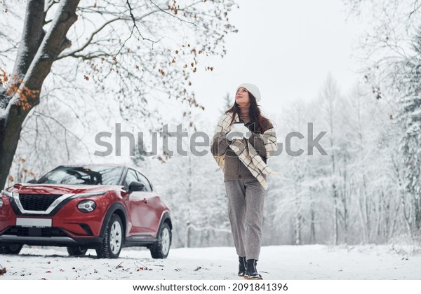 Non urban scene. Beautiful\
young woman is outdoors near her red automobile at winter\
time.