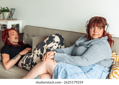 Non-binary person with red hair and mustache is using a laptop while lying on a sofa next to a plus-size woman wearing headphones and listening to music, both enjoying their time at home