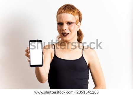 A non-binary redhead in makeup poses in a black T-shirt on a white background while showing a mobile phone with a white screen. Concept of non-binary people, mobile advertising.