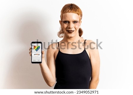 A non-binary redhead poses on a white background in casual clothing showing a mobile phone with a same-sex dating app. Dating concept for gays, application for homosexuals. Application to meet people.