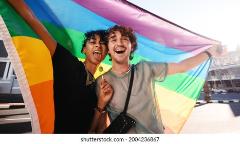 Non-conforming young men celebrating gay pride. Two young gay men smiling cheerfully while raising a rainbow pride flag. Two members of the LGBTQ+ community celebrating at a gay pride parade. - Powered by Shutterstock