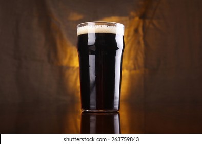 Nonic Pint Of Dark Stout Beer On A Table With A Warm Colored Drapery In The Background