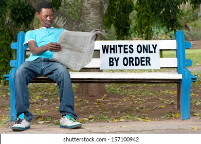 A non-white man sits on a bench in a park reserved for whites. This was commonplace during the apartheid years in South Africa. - Shutterstock ID 157100942