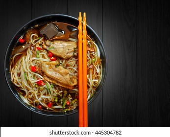 Noodle with chicken in black bowl and orange chopsticks on black wood background. Spicy noodle. Still life. Copy space.