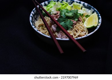 Noodles Served In Bowls With Lemon Wedges, Coriander Garnish And Minced Pork Are Placed On A Black Table.