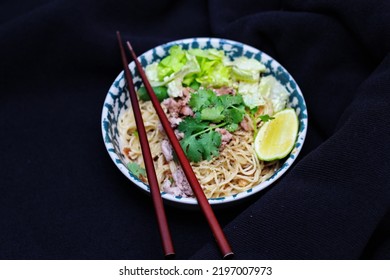 Noodles Served In Bowls With Lemon Wedges, Coriander Garnish And Minced Pork Are Placed On A Black Table.