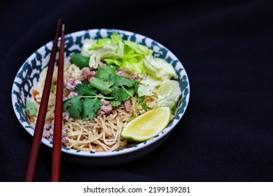 Noodles Served In Bowls With Lemon Wedges, Coriander Garnish And Minced Pork Are Placed On A Black Table.