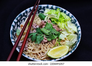 Noodles Served In Bowls With Lemon Wedges, Coriander Garnish And Minced Pork Are Placed On A Black Table.