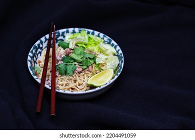 Noodles Served In Bowls With Lemon Wedges, Coriander Garnish And Minced Pork Are Placed On A Black Table.