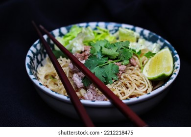 Noodles Served In Bowls With Lemon Wedges, Coriander Garnish And Minced Pork Are Placed On A Black Table.