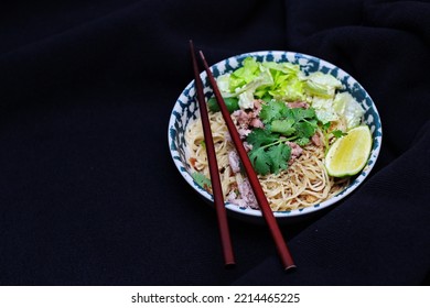 Noodles Served In Bowls With Lemon Wedges, Coriander Garnish And Minced Pork Are Placed On A Black Table.