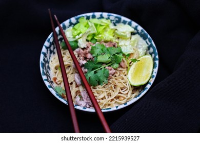 Noodles Served In Bowls With Lemon Wedges, Coriander Garnish And Minced Pork Are Placed On A Black Table.