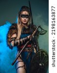 Nordic huntress woman with strup back longbow holding shield and axe against teal background with smoke.