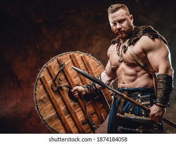 Nordic warlike barbarian with muscular build and naked torso holding shield and spear in dark background.