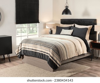 A Nordic-style bedroom with a black upholstered headboard, pillows, a striped quilt in beige and black tones, side tables, a window with a garden view, pendant light, all atop a beige area rug.