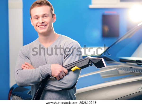 Normal Saturday of car\
owner. Closeup of handsome smiling young man standing by his luxury\
car with his hands crossed while holding a car vacuum cleaner car\
service
