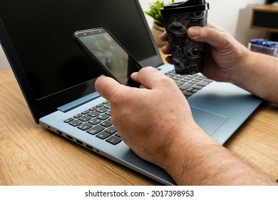 Normal view of a table with a man consulting and checking his cell phone while working on his notebook at home. Technology and communications concept