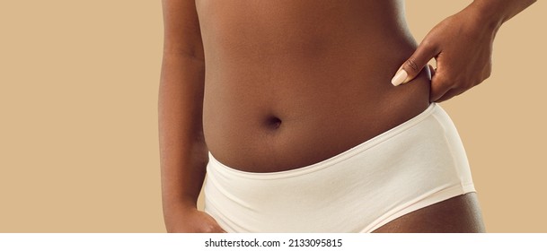Normalizing body flaws, accepting yourself and being confident in your imperfection: Plump young black woman in underwear, with a cute naked belly button, pinching some body fat on her side, close up