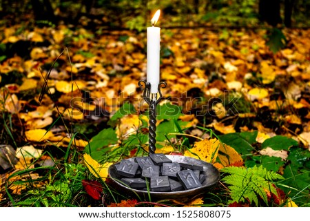 Norse runes in a forged bowl with a candle on the background of autumn forest foliage.
