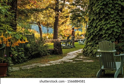 North Hatley, PQ, Canada - September 4, 2014 : A Youthful Baby Boomer Woman Stands Beside An Adirondack Style Chair In A Lush Garden Framed By A Wall Of Vines At Sunset