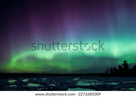 Northern Lights Brilliant Aurora Sky erupt over a lake in Minnesota in the dark sky overhead shining a rainbow of light and colors over the forests and frozen water and ice