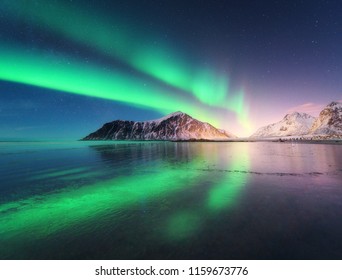 Northern lights in Lofoten islands, Norway. Green Aurora borealis. Starry sky with polar lights. Night winter landscape with aurora, sea with sky reflection, rocks, beach and snowy mountains. Travel - Shutterstock ID 1159673776