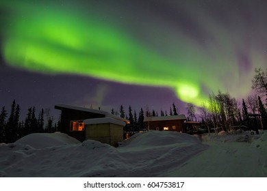 Northern lights over snow covered road leading up to cabins.