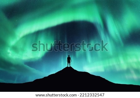 Northern lights and silhouette of standing man on the top of mountain. Aurora borealis and happy guy. Sky with stars and green polar lights. Colorful night landscape with aurora background. Concept