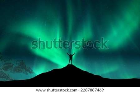 Northern lights and silhouette of standing man with raised up arms on the mountain peak in Norway. Aurora borealis and happy man. Sky with stars and polar lights. Night landscape with aurora. Concept