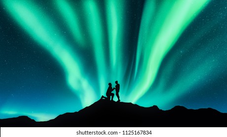 Northern Lights And Silhouettes Of A Man Making Marriage Proposal To His Girlfriend On The Hill. Landscape With Night Starry Sky, Aurora Borealis, Couple. People. Lovers And Polar Lights. Travel