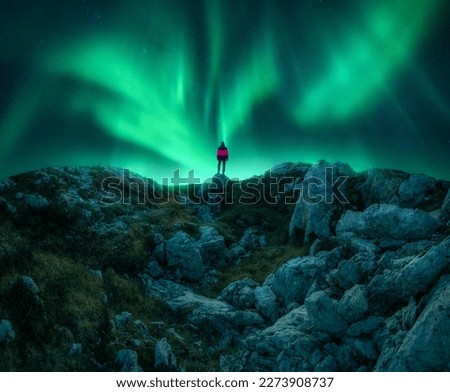 Northern lights and young woman on mountain peak at night. Aurora borealis, stones and silhouette of alone girl on mountain trail. Landscape with polar lights. Starry sky with bright aurora. Travel