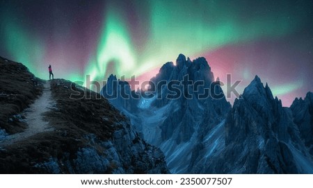 Northern lights and young woman on mountain peak at night. Green and violet aurora borealis and silhouette of alone girl on mountain trail. Landscape with polar lights. Starry sky with bright aurora