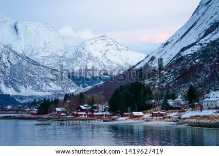 Norway, the village Ornes in December at noon. Typical wooden houses on the coastline an in the background snow covered mountains.