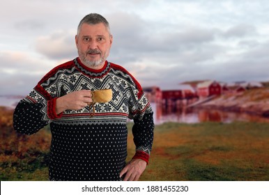 A Norwegian With A Beard Drinks Coffee From A Wooden Cup. The Older Man Wears A Typical Red And Black Norwegian Sweater With Nordic Patterns. In The Background Is A Bay With Wooden Houses.