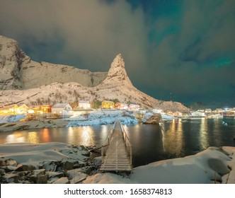 Norwegian Fishing Village With Northern Lights And Stars In Reine City, Lofoten Islands, Norway, Europe. White Snowy Mountain Hills And Trees At Night, Nature Landscape Background In Winter Season.