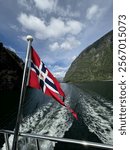 Norwegian flag waving on a boat, with fjord waters and towering green cliffs in the background under a partly cloudy blue sky.