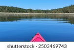 The nose of a red kayak in the foreground floating on calm blue water, with shoreline and trees in the distance. 