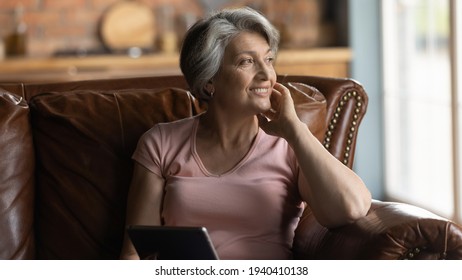 Nostalgia. Peaceful Smiling Elderly Aged Female Grandma Sit On Comfy Couch Hold Tablet Dream Distracted From Gadget Screen. Pensive Older Hispanic Woman Rest Of Pad Computer Lost In Positive Thoughts